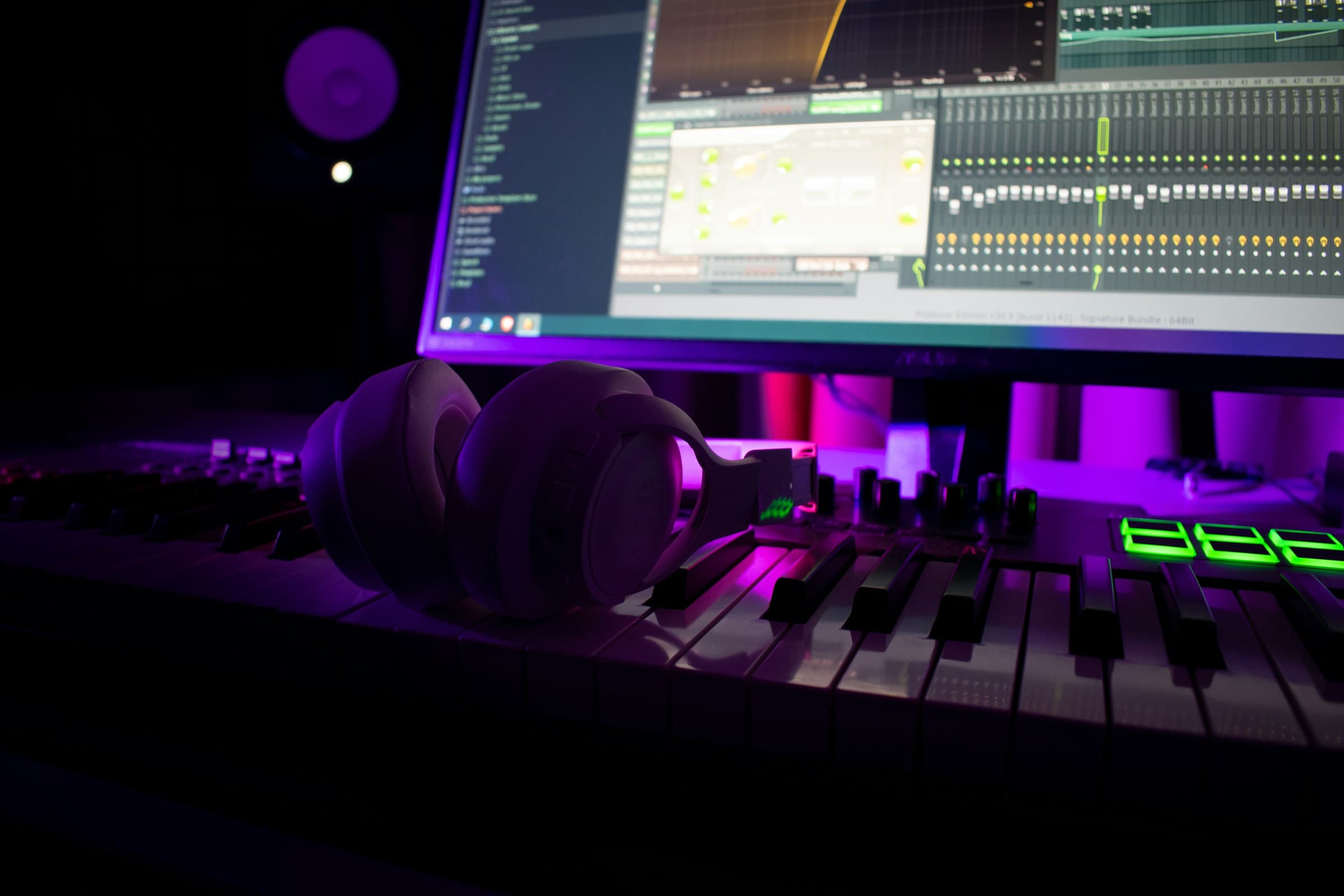 Headphones resting on a keyboard in front of a music-production workstation lit in purple and green
