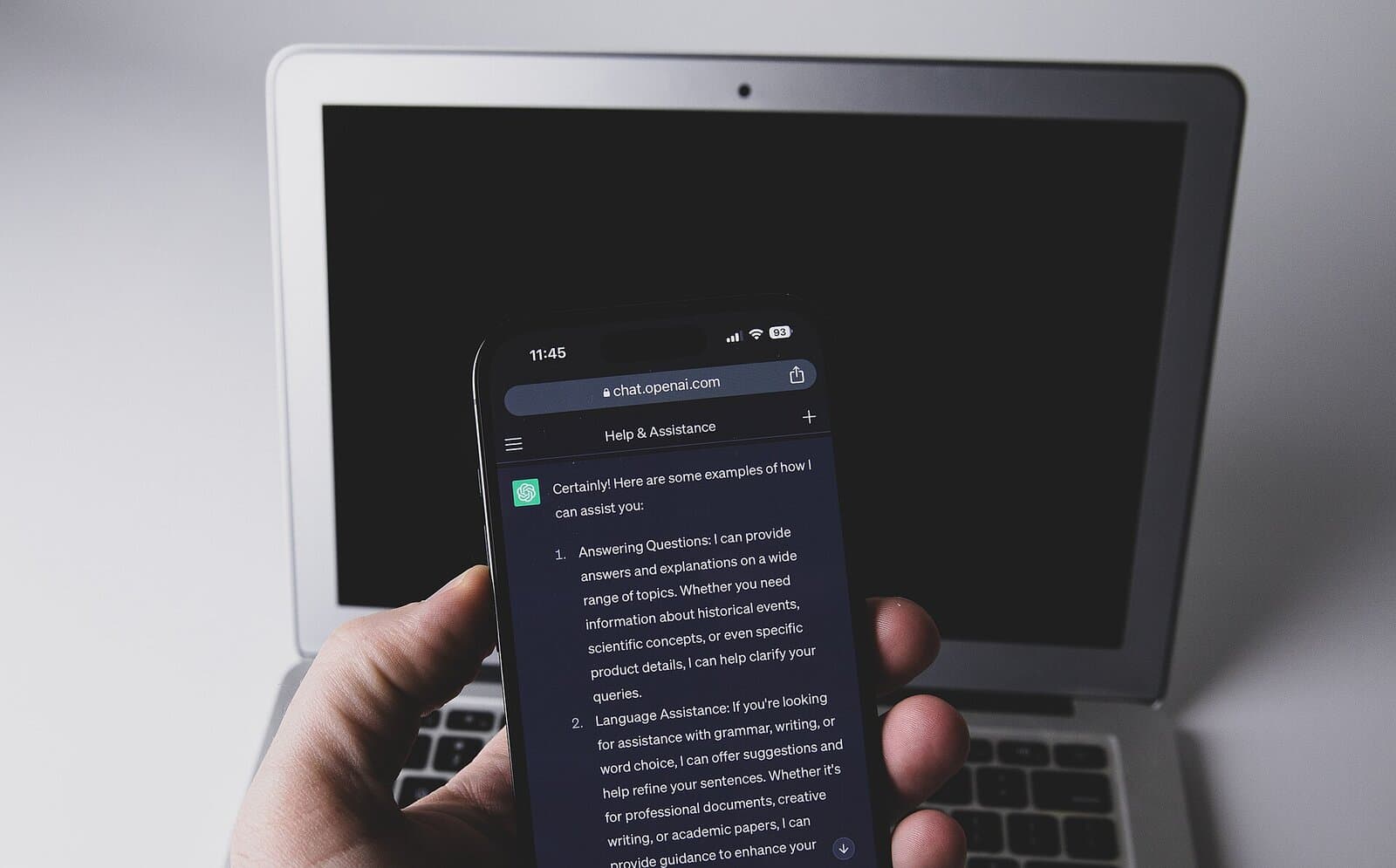 A hand holds a phone with ChatGPT open in front of a laptop