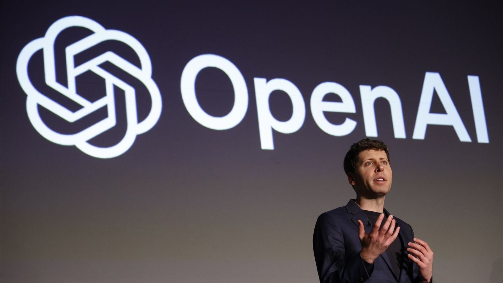 Sam Altman speaks on stage beneath a giant OpenAI logo