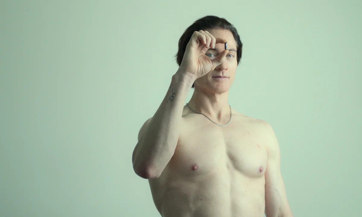 Bryan Johnson, shirtless, holds a pill between his fingers in front of his face