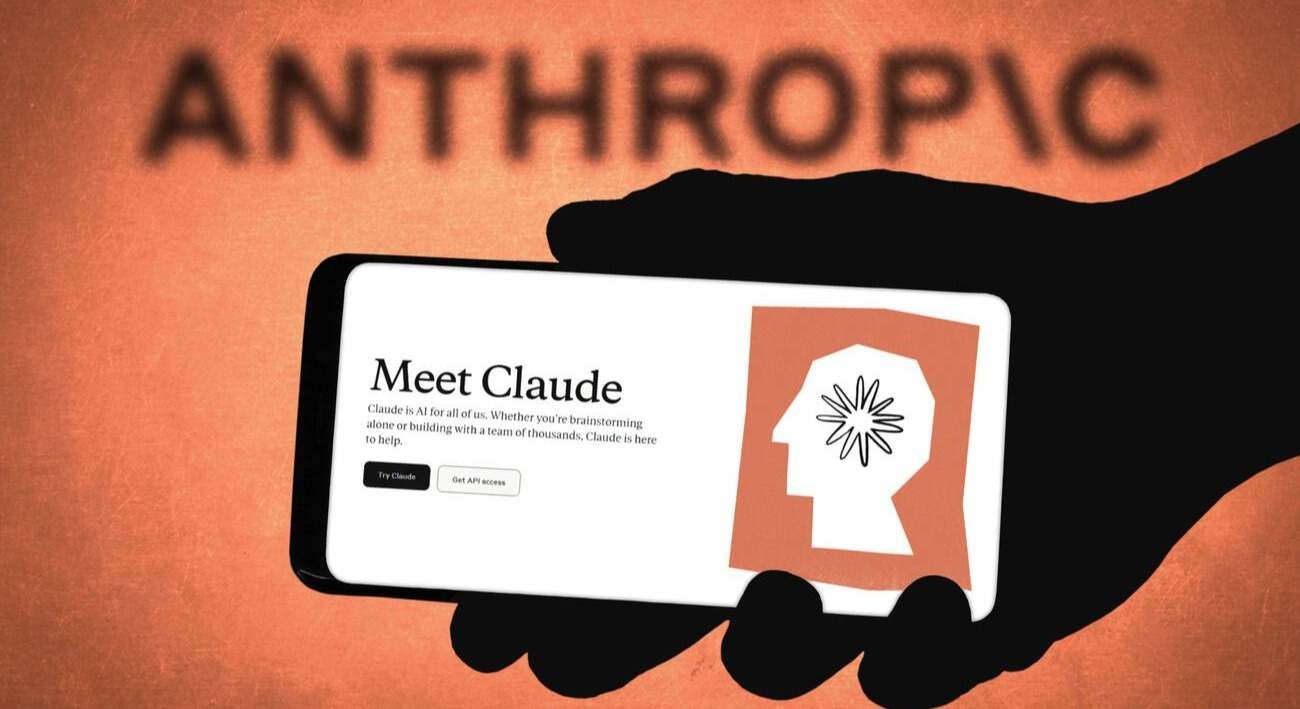 A hand holds a phone showing the Meet Claude page in front of the Anthropic logo