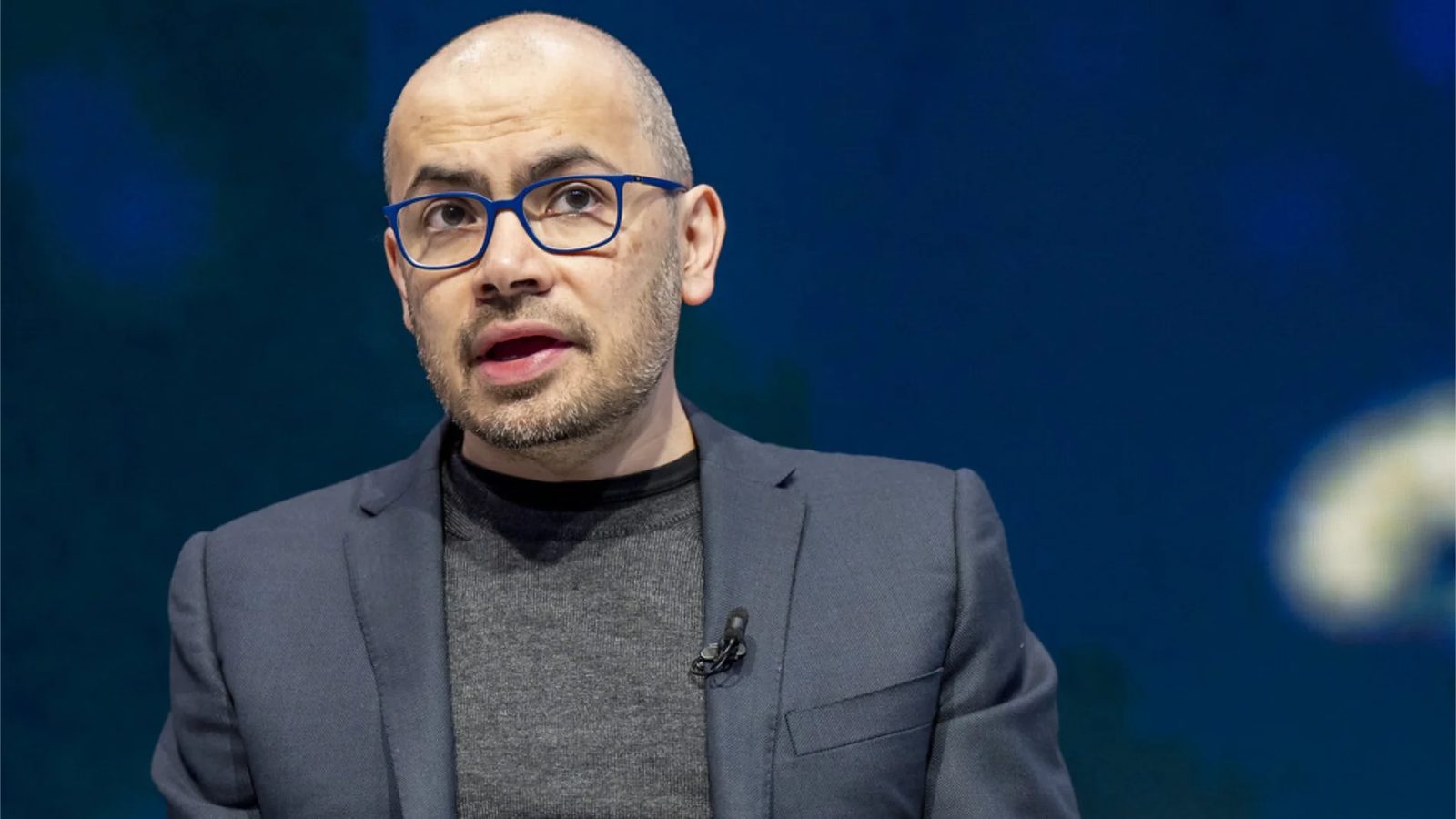 Demis Hassabis, wearing blue glasses, speaks on stage against a dark background