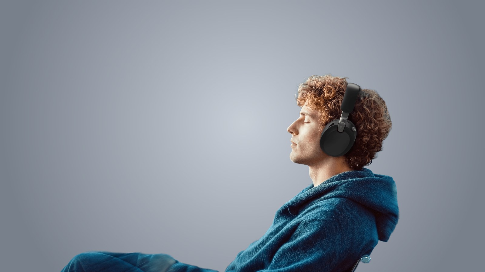 A young man with curly hair and his eyes closed wearing the black MSI Maestro 500 Wireless, leaning back under a blue blanket against a blue-gray backdrop
