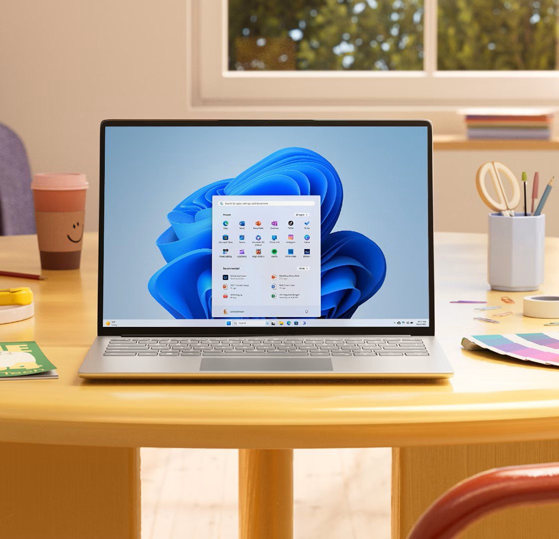 Portátil con Windows 11 y el menú Inicio abierto sobre una mesa de madera junto a una ventana
