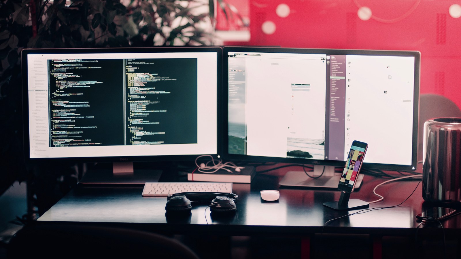 A programming workstation with two monitors full of code and a phone on a stand, lit in red tones