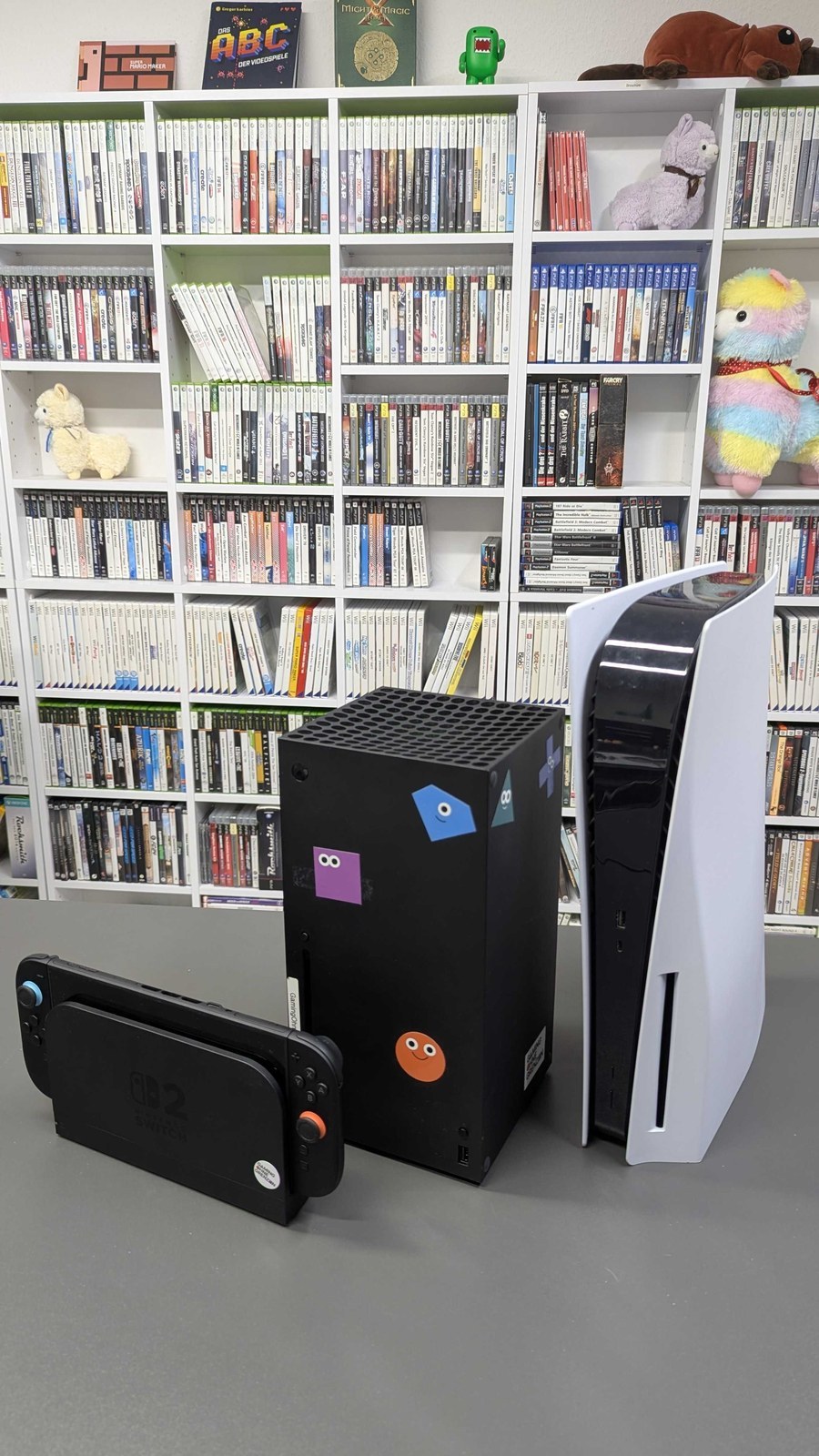 A Nintendo Switch 2, an Xbox Series X and a PlayStation 5 on a table in front of shelves of games