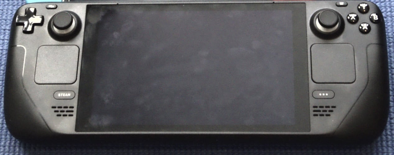 Vista frontal de una Steam Deck LCD con la pantalla apagada sobre una superficie azul