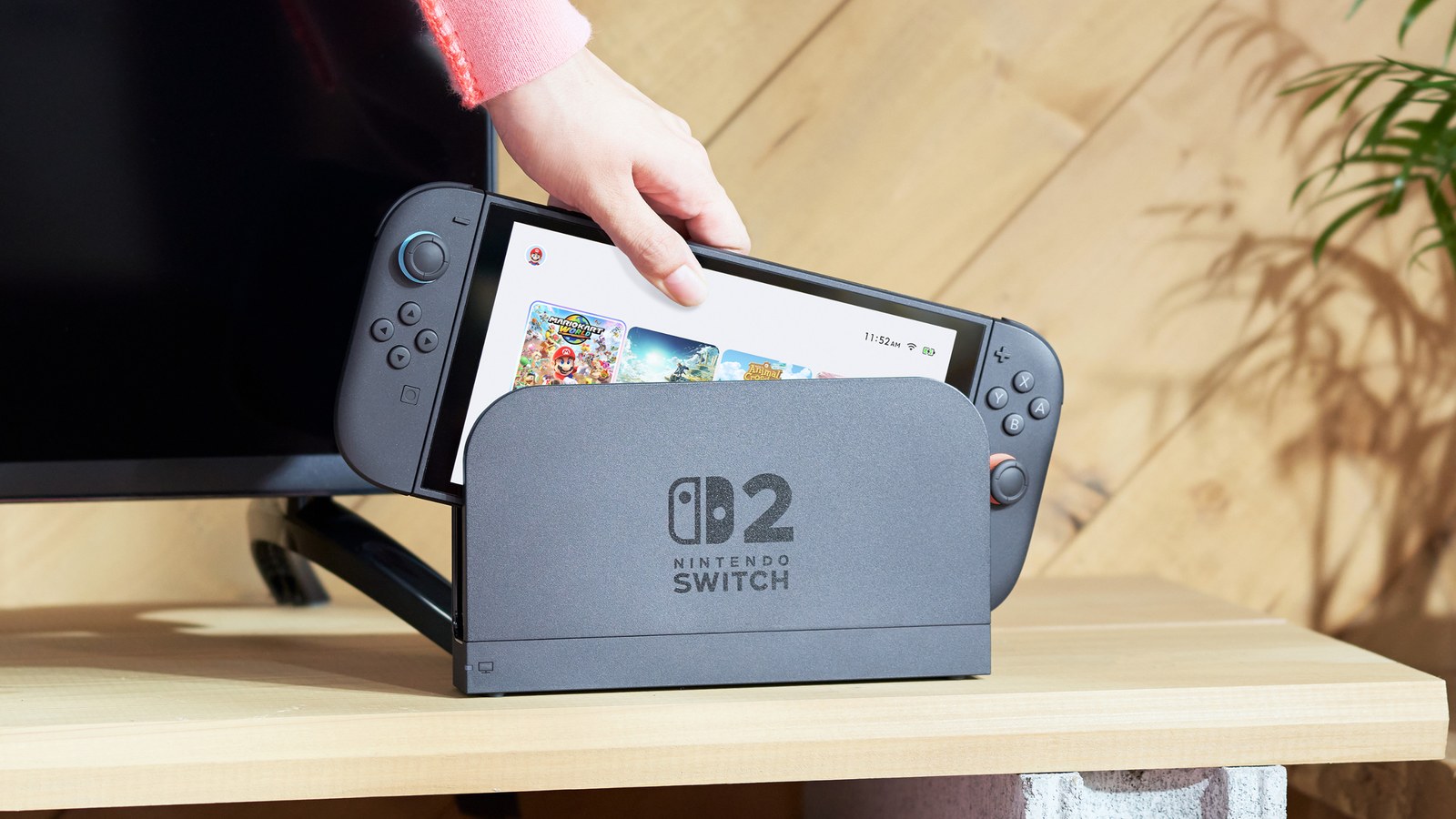 Una mano coloca una Nintendo Switch 2 en su base, sobre un mueble de madera junto al televisor