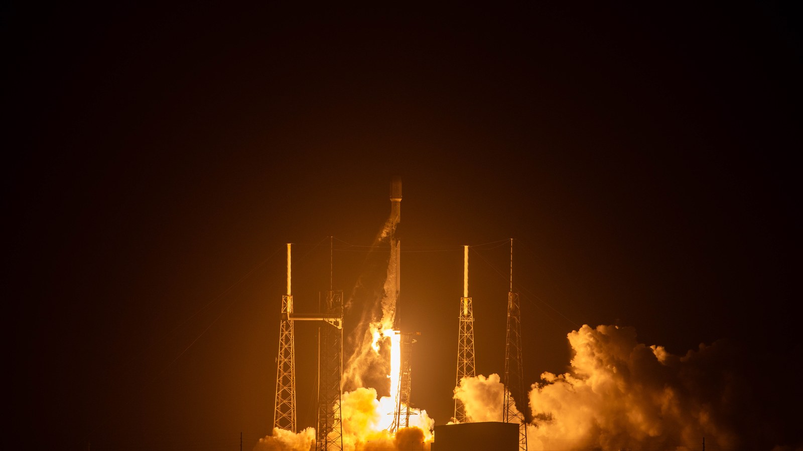 A SpaceX Falcon 9 rocket lifts off at night carrying Starlink satellites, lit orange between the launch pad towers on October 7, 2025