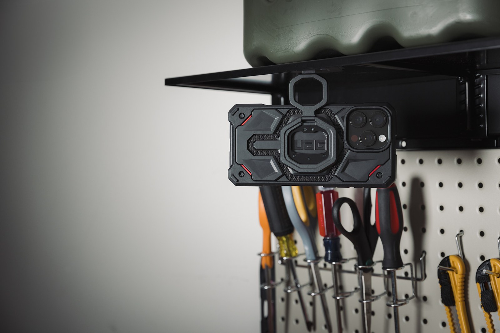 A phone hangs beneath a metal workshop shelf, held only by the outer magnetic face of the Monarch Mag2