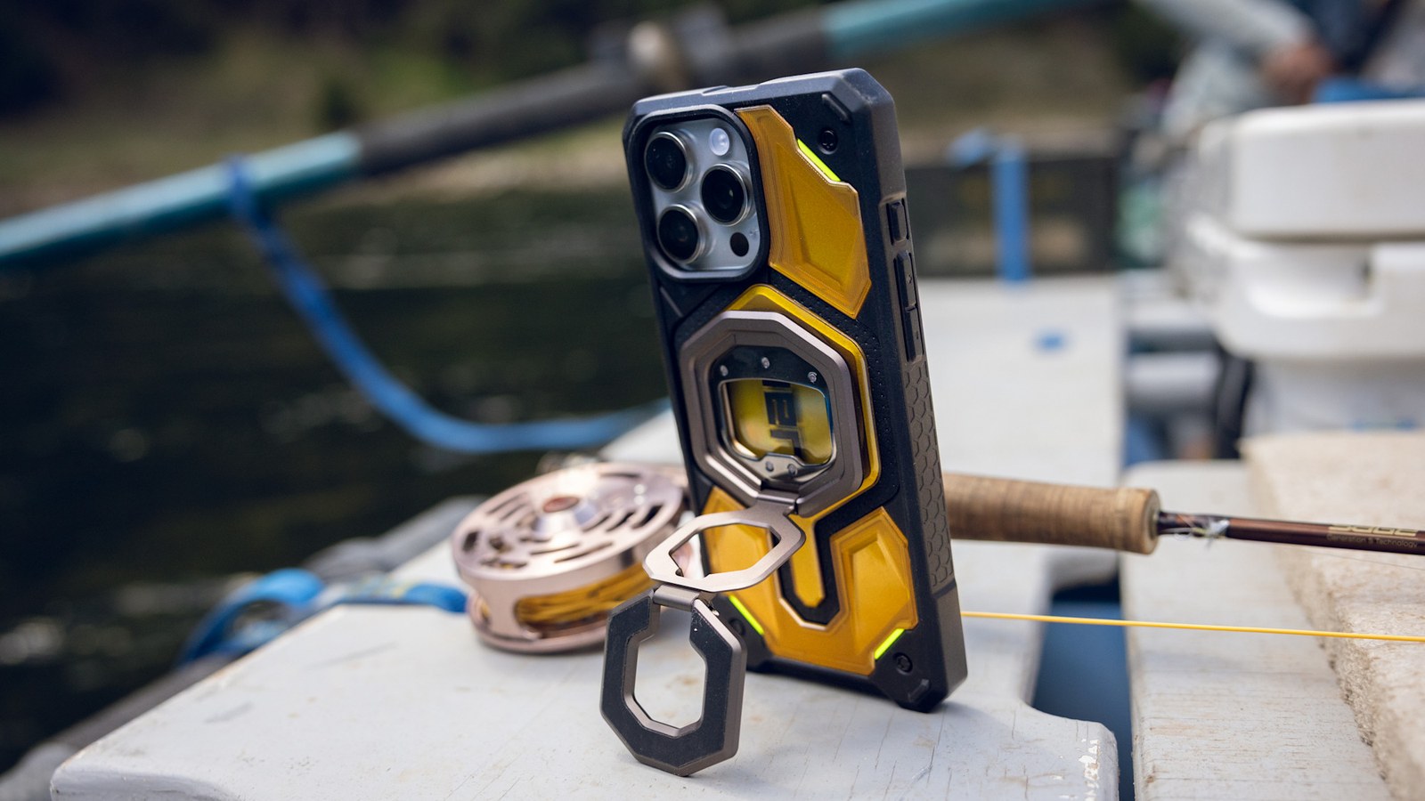iPhone con funda amarilla de UAG apoyado de pie en la cubierta de una barca gracias a la pata del anillo Monarch Mag2, junto a una caña de pescar