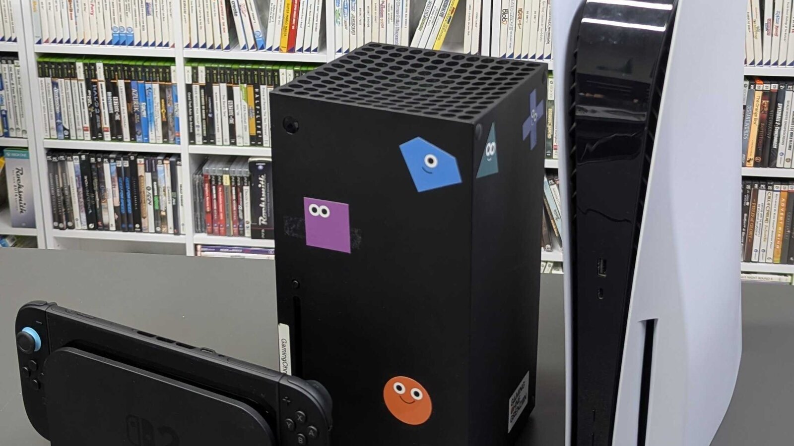 A Nintendo Switch 2 with its controllers, a black Xbox Series X covered in stickers and an upright white PS5 on a gray table in front of shelves full of games